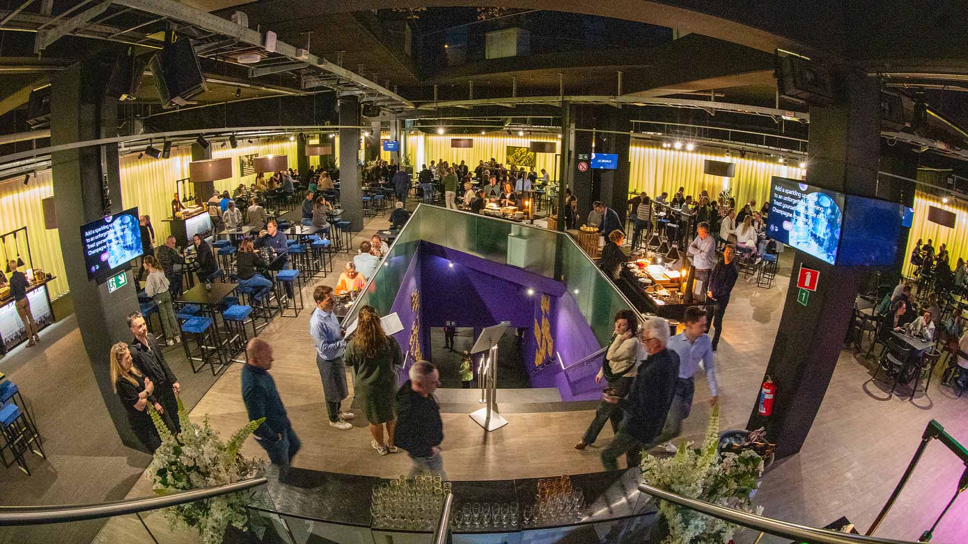 Een panoramisch bovenaanzicht van het ruime Hospitality Center, gevuld met bezoekers die rondlopen en plaatsnemen aan tafels, met een opvallende paarse trap in het midden.
