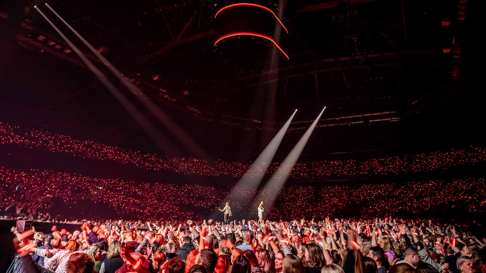 Koen and Kris Wauters of Clouseau are performing in the center of the AFAS Dome. Many fans on the floor have their hands in the air, and red lights are visible throughout the stands