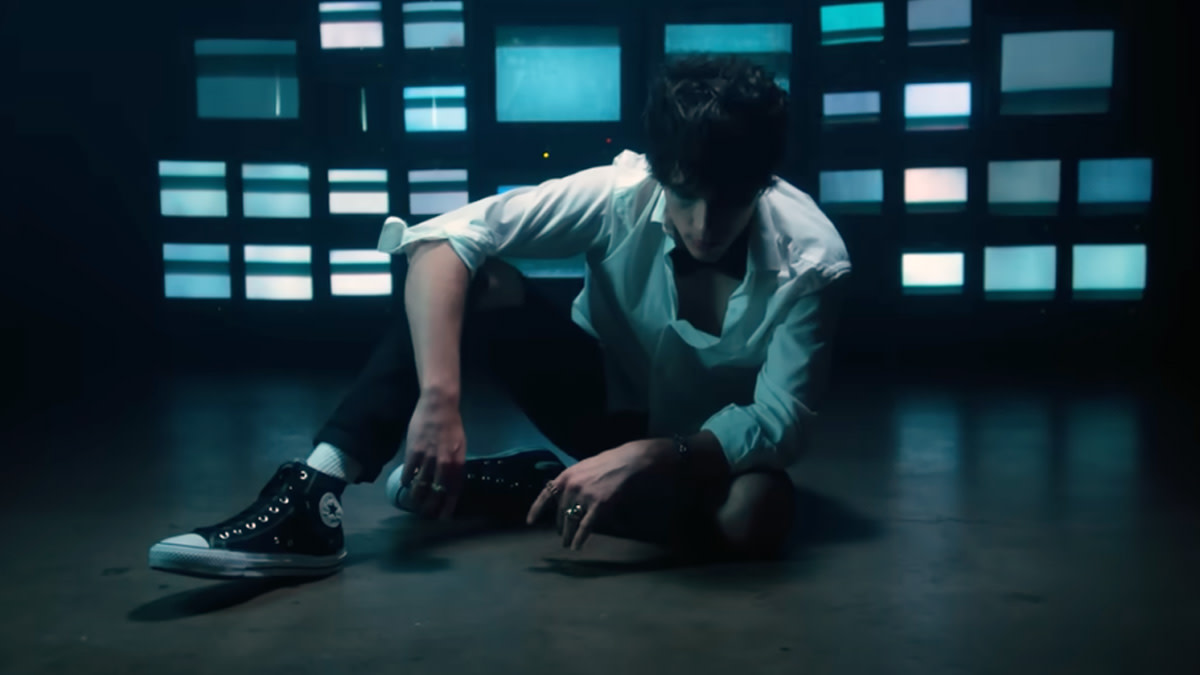 The singer from Weathers is sitting on the floor in front of a wall with television screens.