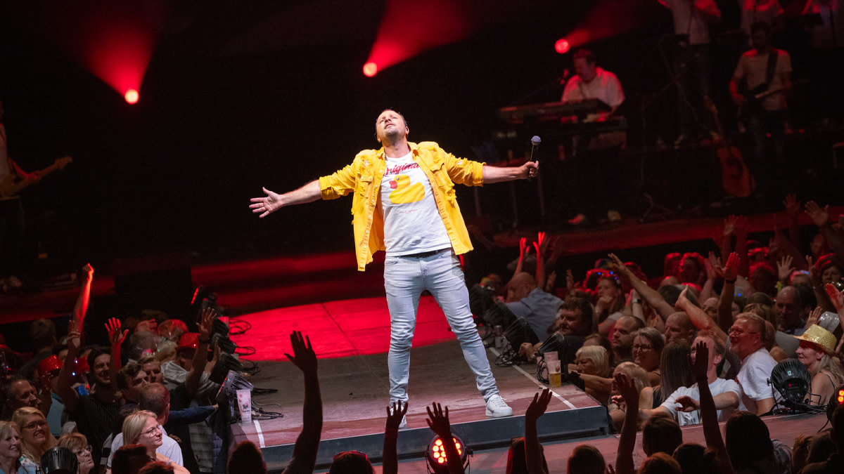Johan Veugelers in een witte t-shirt en geel jasje op het podium omringd door fans.