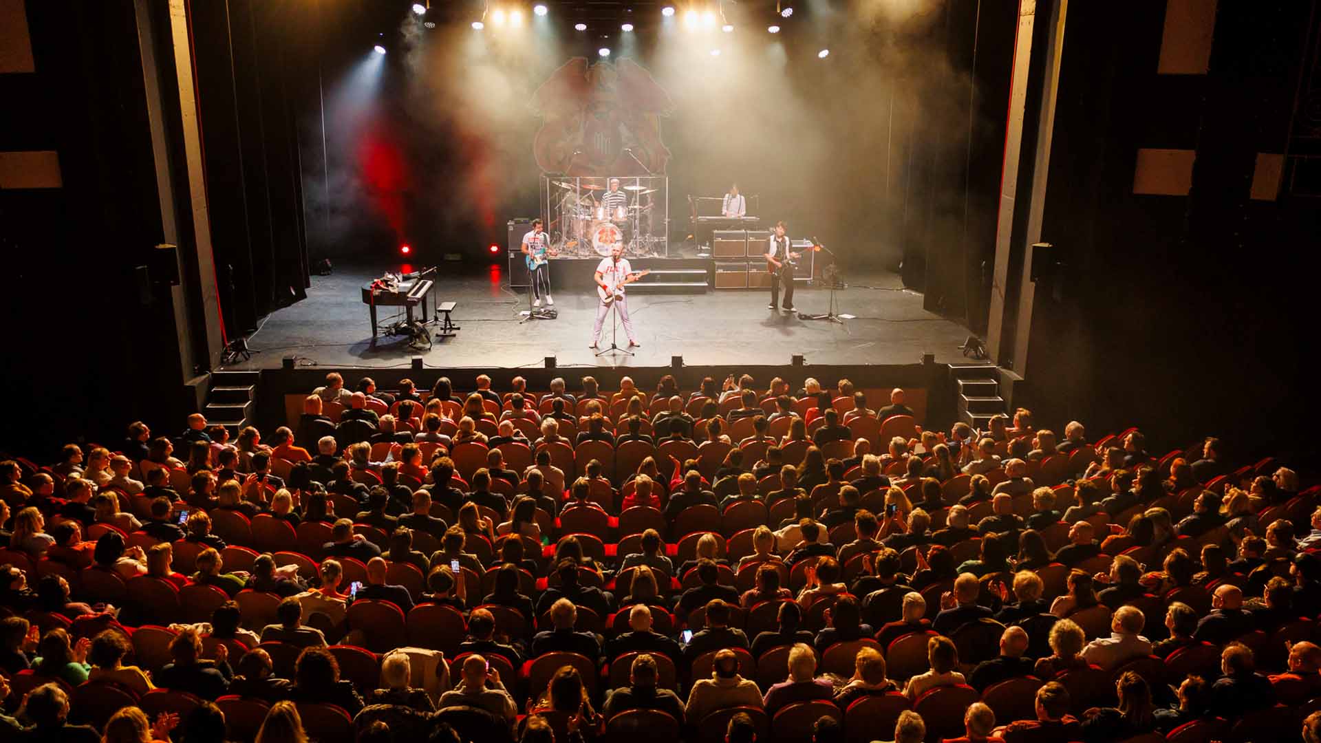 Op het podium staat de band Mother Mercury. Het publiek kijkt toe vanuit de zaal.