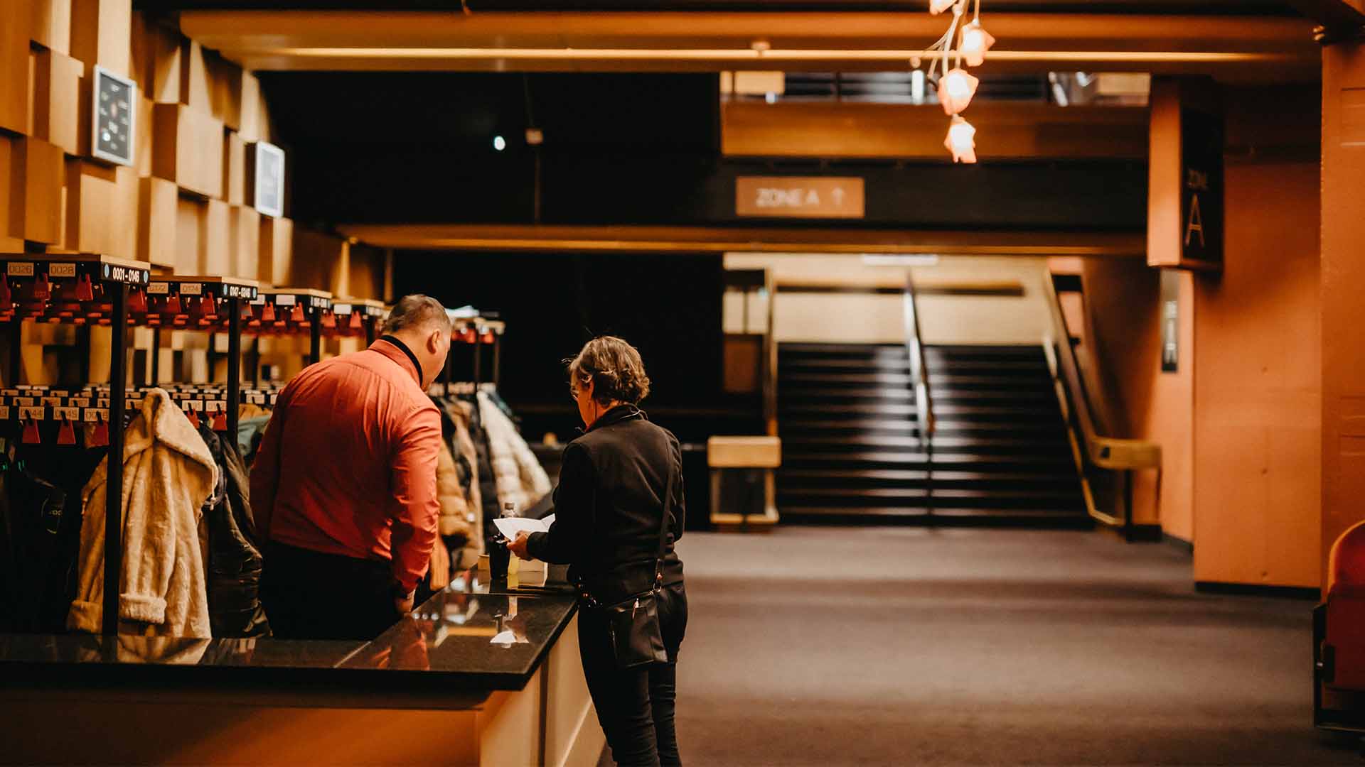 Two people at the cloakroom in Stadsschouwburg Antwerpen. Coats hang on the racks in the background, along with a staircase leading to the auditorium.