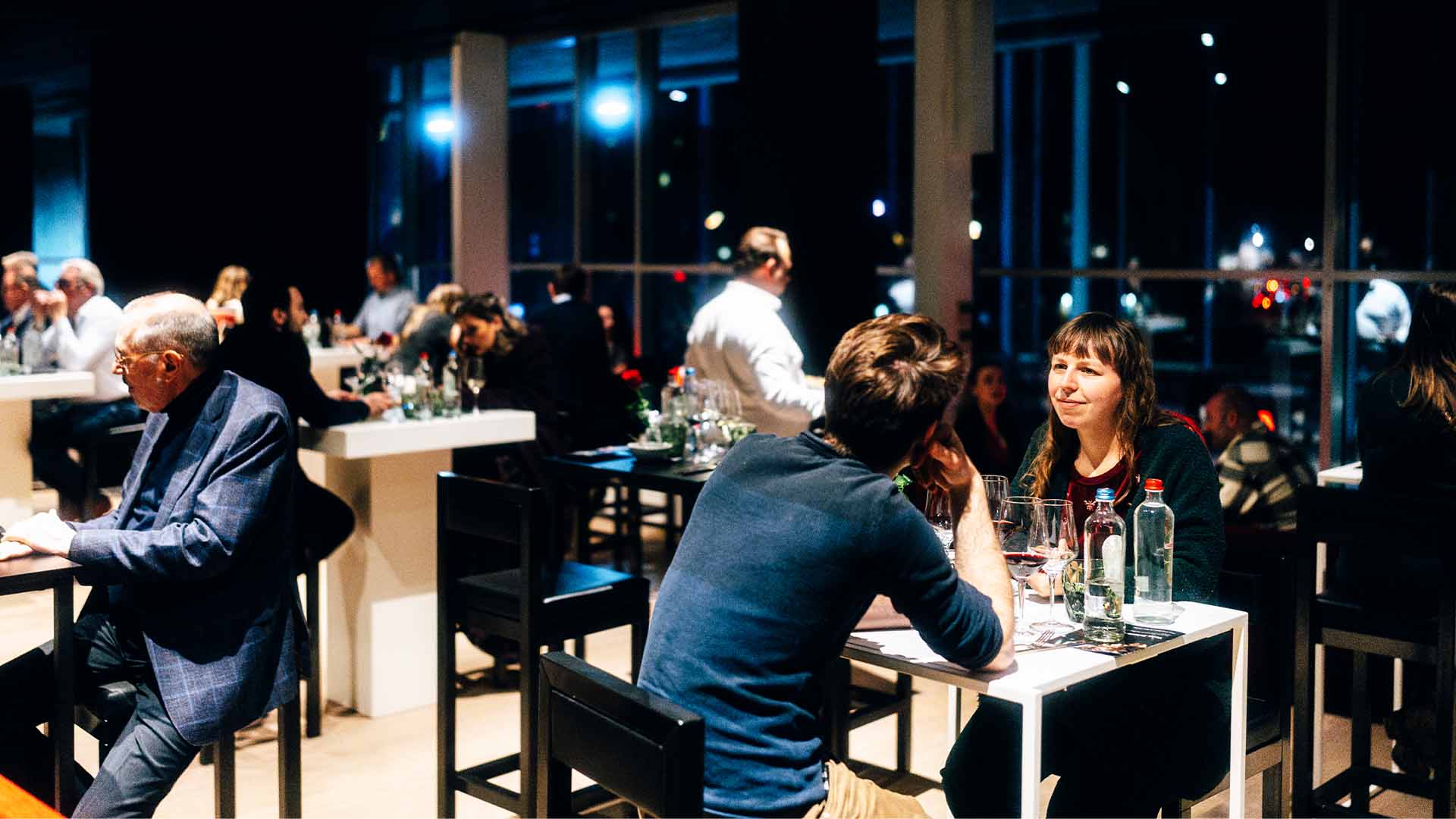 Een koppel zit aan een hoge tafel in een sfeervolle evenementenruimte, met andere gasten op de achtergrond.