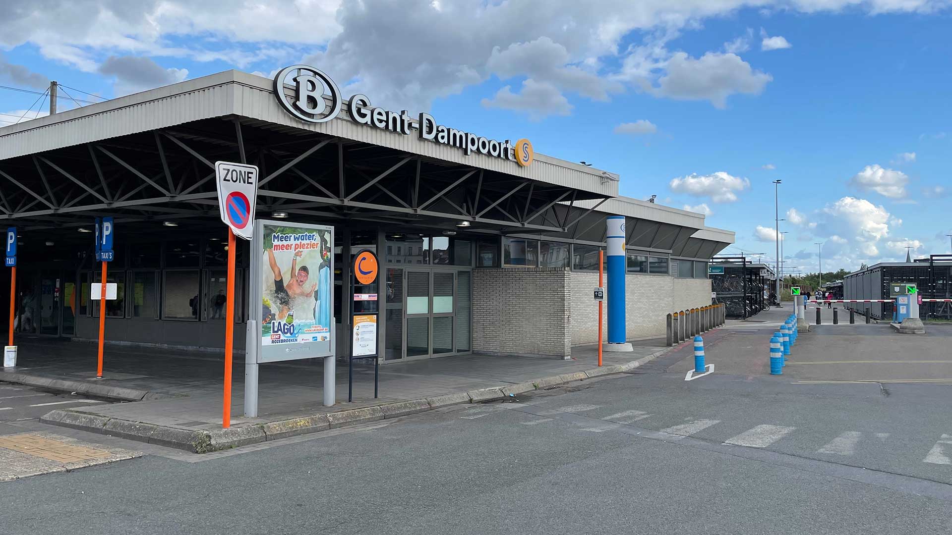 Station Gent-Dampoort met de hoofdingang, stationsgebouw en zichtbare stationssignalisatie, gelegen naast parkeerplaatsen onder een gedeeltelijk bewolkte hemel.