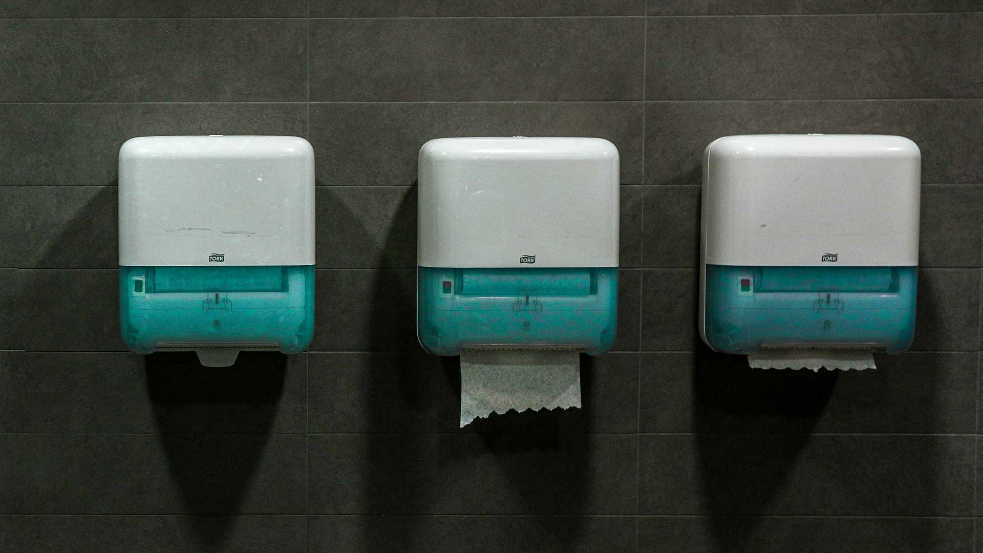 Trois distributeurs de serviettes en papier Tork blancs et cyan sont suspendus côte à côte sur un mur de carreaux gris. Dans le distributeur du milieu, une serviette est visible. Dans le distributeur de droite, un petit morceau de papier est visible.