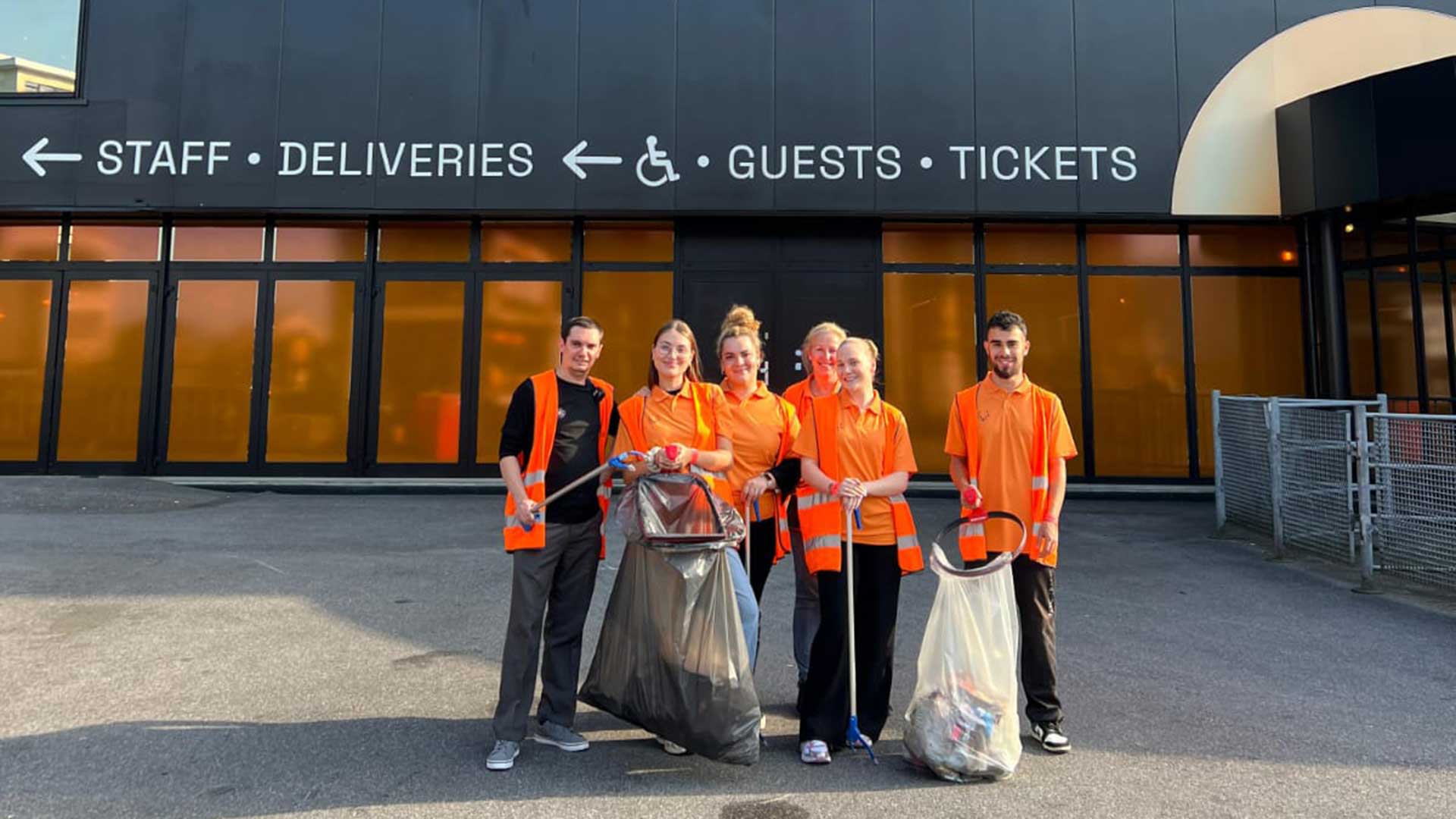 Six personnes en gilets et chemises orange sourient devant Forest National. Elles tiennent des pinces et des sacs remplis de déchets ramassés lors d’une action de nettoyage.
