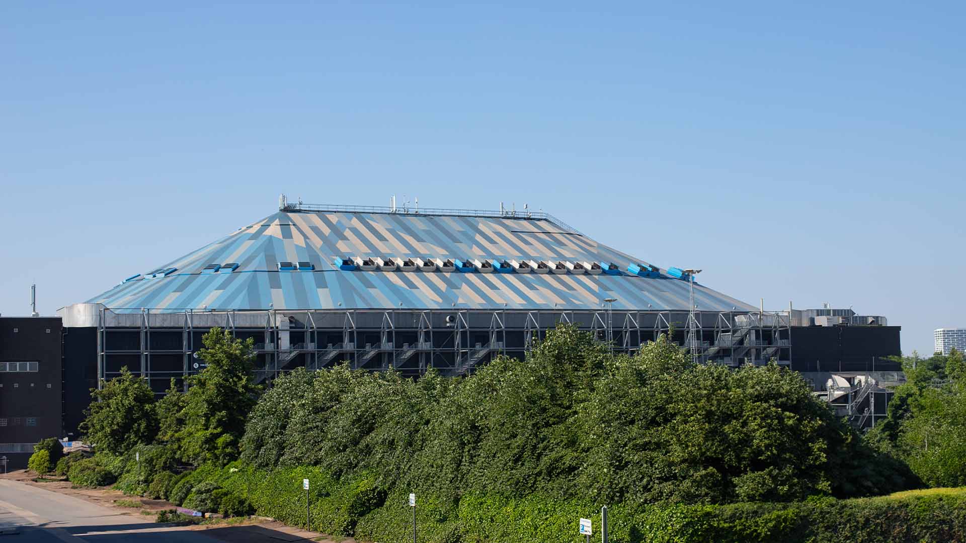 Exterior of the AFAS Dome in Antwerp featuring its distinctive blue-and-white roof, viewed from the side and surrounded by greenery under a clear sky.