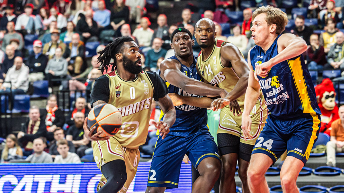Een speler van Antwerp Giants dribbelt met de bal richting de basket tijdens een aanval, terwijl meerdere verdedigers hem proberen te stoppen. Rondom hen zit het publiek in de tribunes aandachtig te kijken naar de actie op het veld.