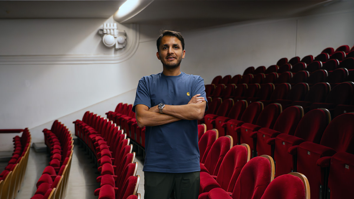 Comedian Mohsin Abbas staat met gekruiste armen tussen de rode theaterstoelen en kijkt glimlachend in de camera.