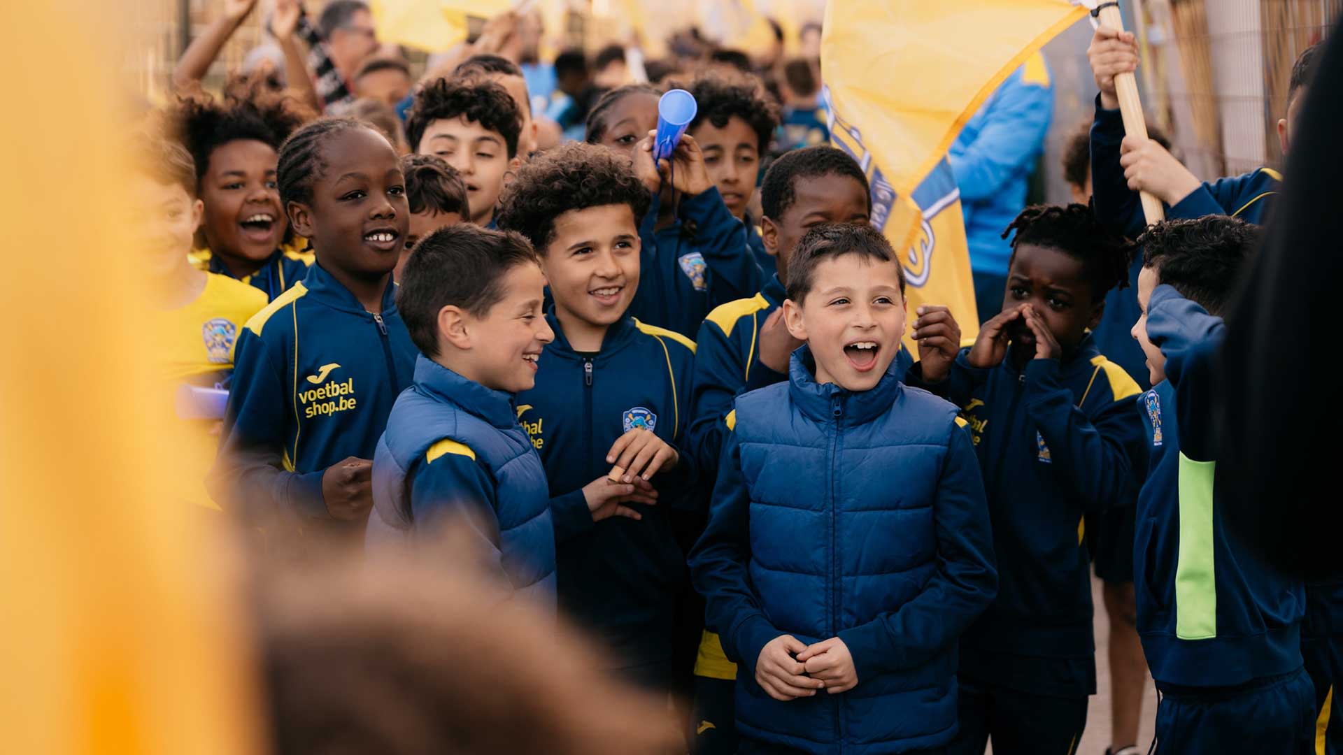 Een groep jonge spelers van City Pirates in blauwe trainingspakken en gewatteerde jassen, verzameld in een rij met gele en blauwe vlaggen tijdens een clubactiviteit.