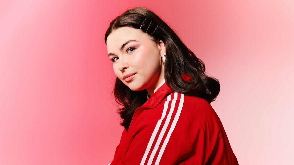Ruby XX met lang zwart haar in een rood trainingspak tegen een roze achtergrond.