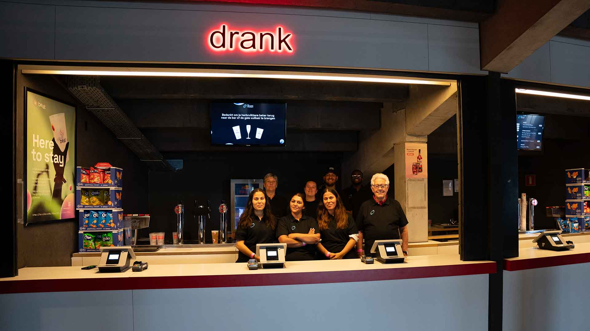 Een team van zeven mensen, die in zwarte shirts achter een bar staan. Boven de bar is het woord ‘Drank’ in rode neonletters te zien. Op de bar staan vier pinautomaten en op de achtergrond zijn stapels chips en enkele tapkranen te zien.