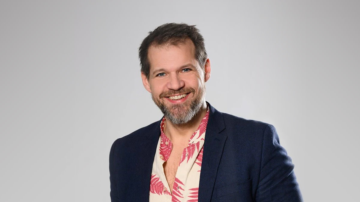 René Van Kooten glimlacht naar de camera, gekleed in een donkerblauwe blazer en een hemd met rood-witte print.