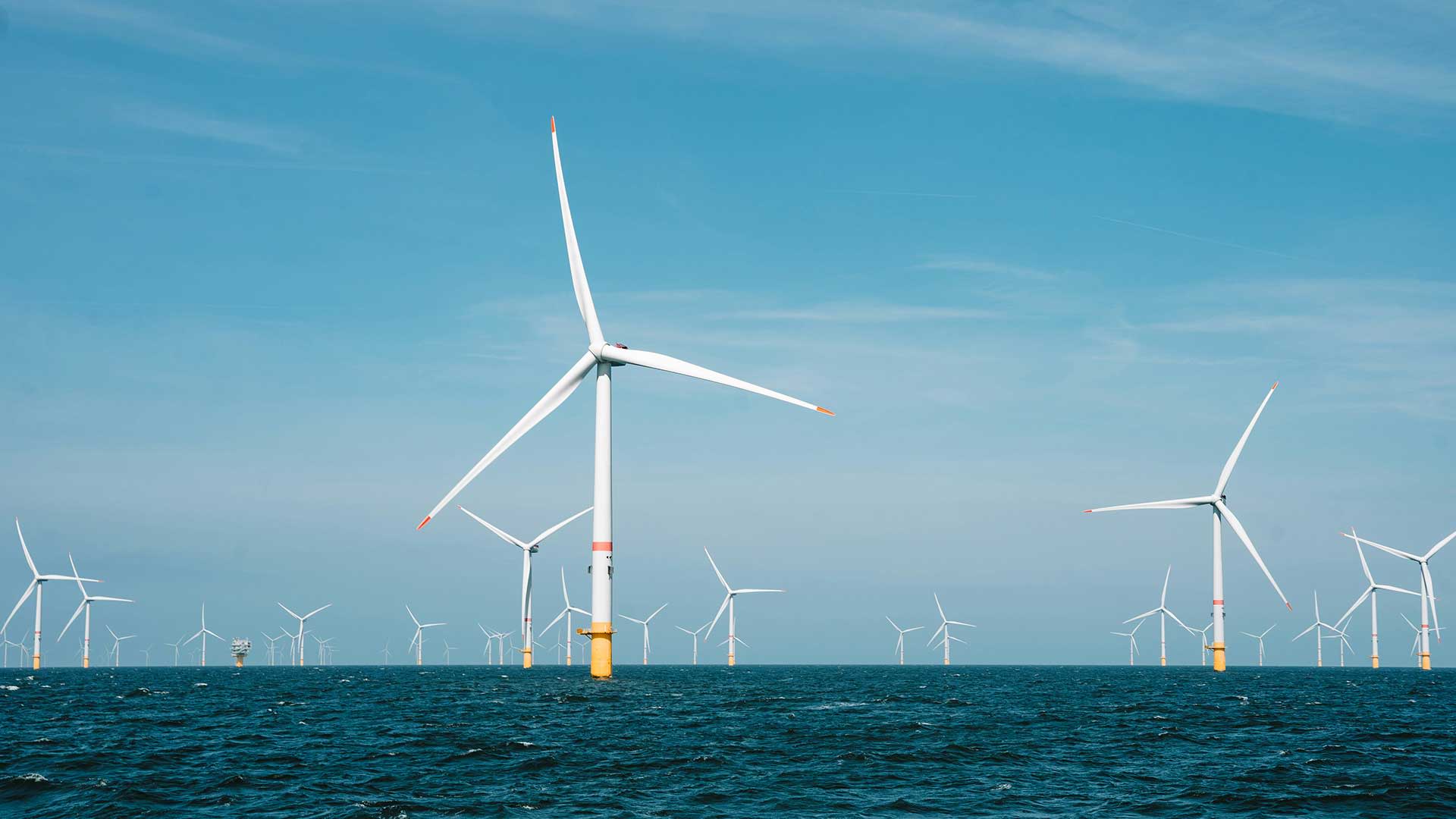 Een offshore windmolenpark onder een heldere blauwe lucht, met talloze windturbines die uit het diepblauwe water oprijzen en hun witte wieken draaien.