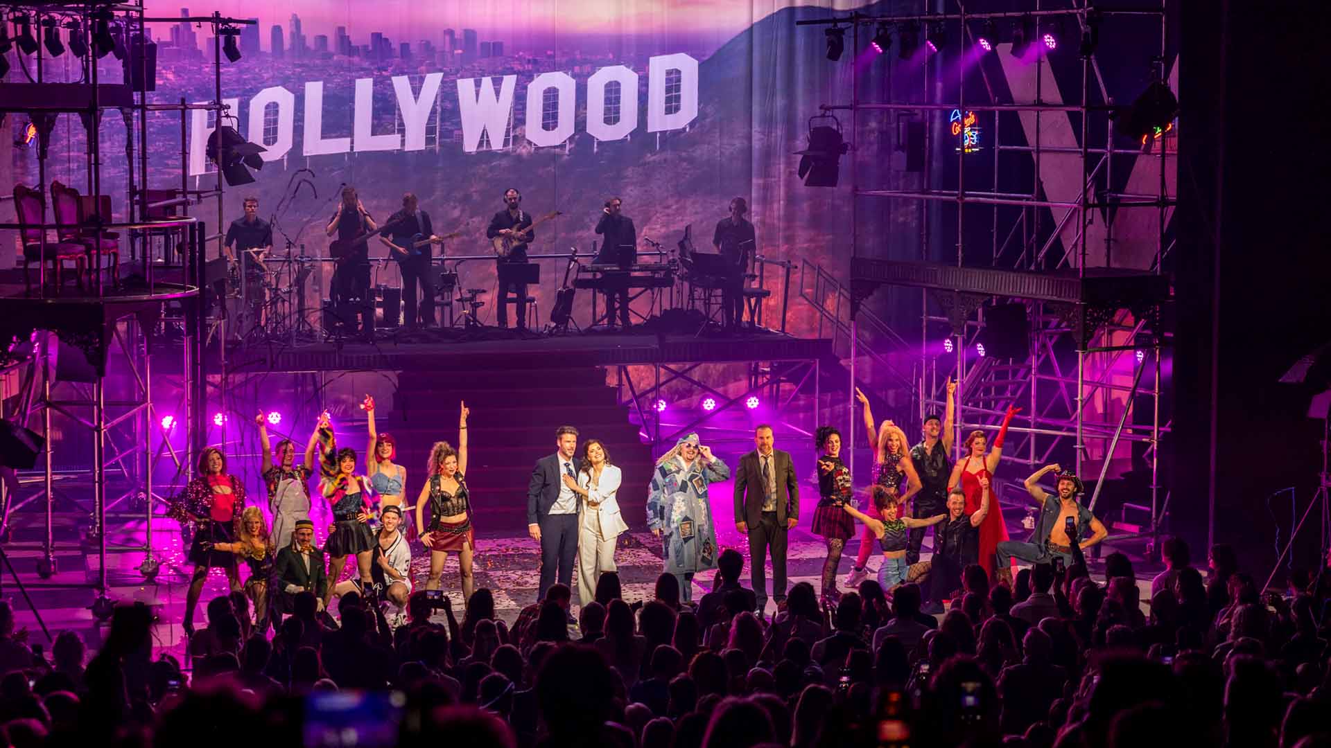 De cast van de musical 'Pretty Woman' staat op het podium. Op de achtergrond speelt de band muziek en boven hen hangen de witte letters van HOLLYWOOD.