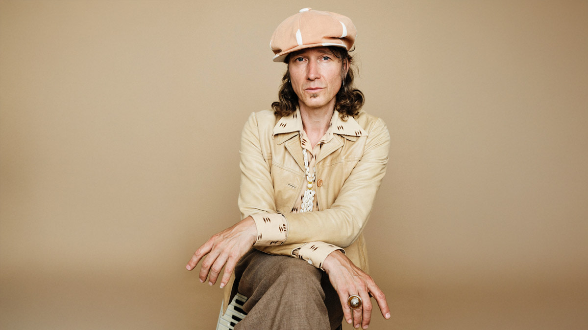 Portret van Stef Kamil Carlens die zittend poseert tegen een beige achtergrond. Hij draagt een lichte pet, een beige jas en bruine broek, met lange krullen zichtbaar onder zijn pet en een opvallende ring aan zijn hand.