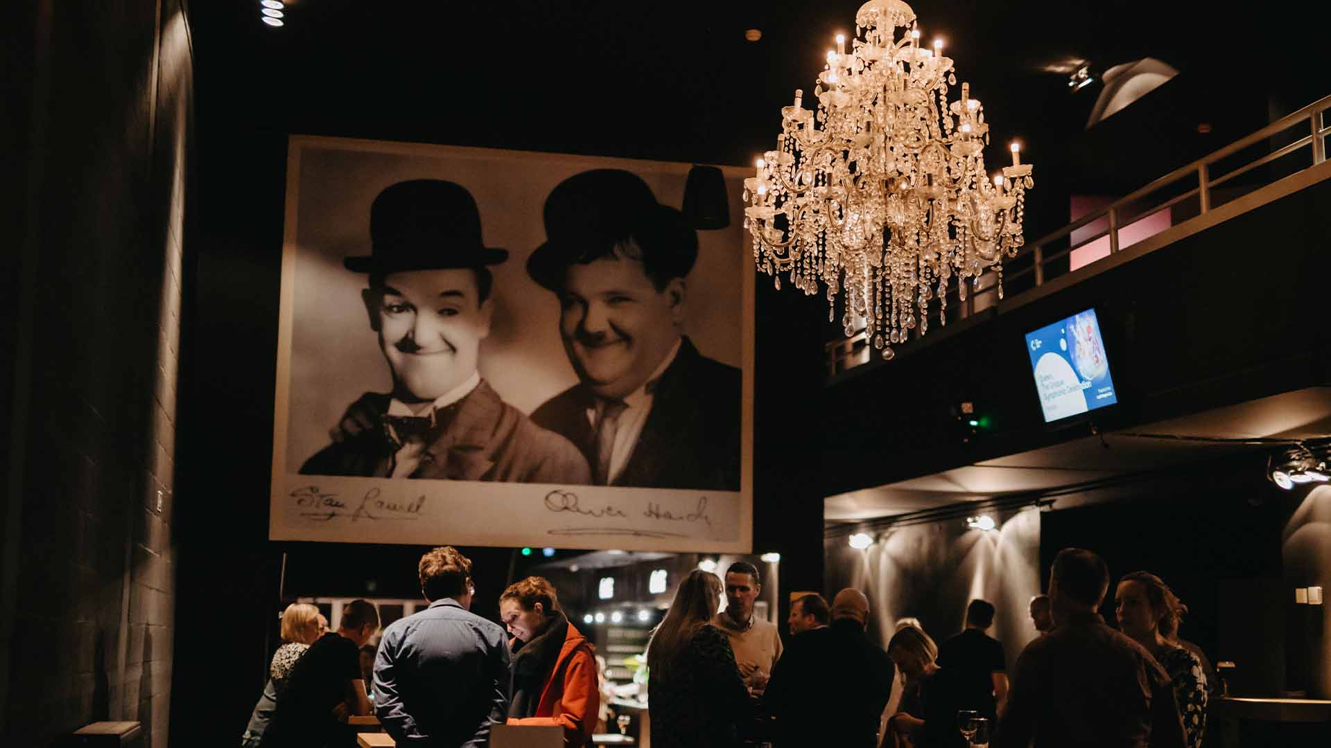 In de Koepelbar staan enkele mensen aan verschillende tafeltjes te praten. Aan het plafond hangt een grote kroonluchter en aan de muur een grote gesigneerde poster van Laurel & Hardy.