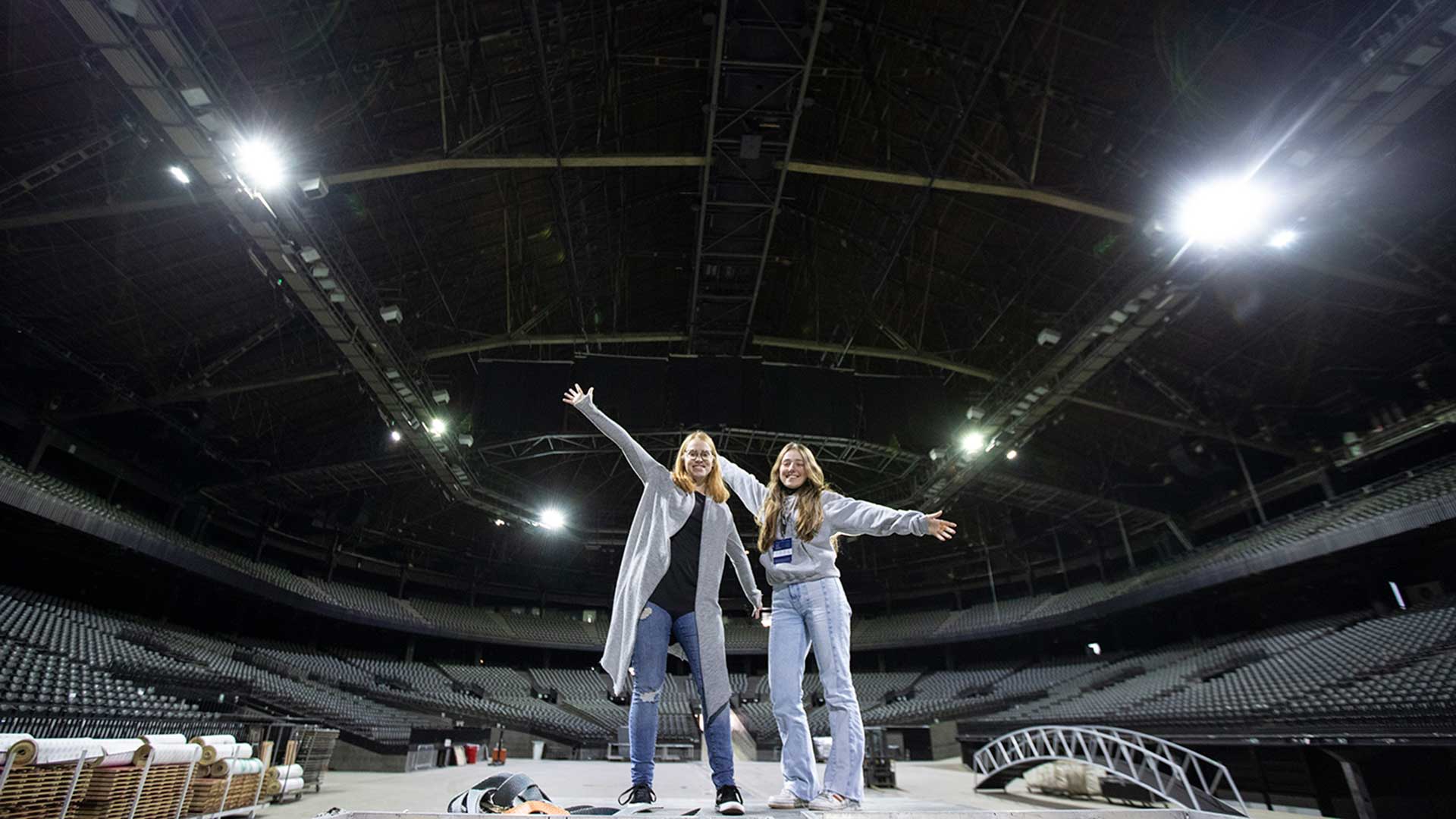 Deux filles souriantes se tiennent les bras écartés sur une scène de l’AFAS Dome. La salle est vide et les gradins sont plongés dans l’obscurité, tandis que la scène et les filles sont éclairées par des lumières vives.
