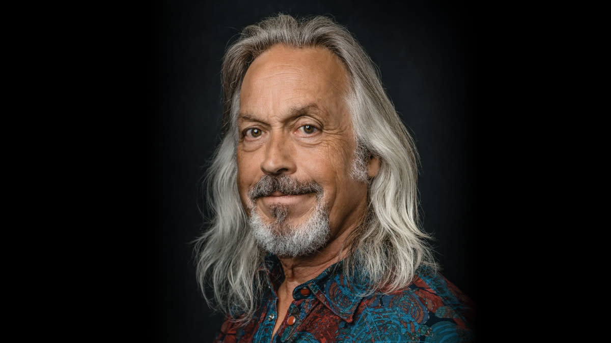 Jim Lauderdale met lange grijze haren en een kleurrijk hemd tegen een donkere achtergrond.