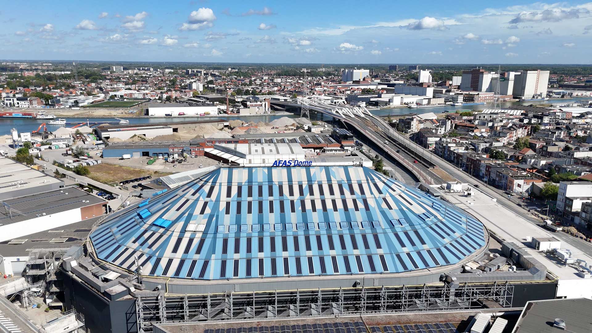 Luchtfoto van de AFAS Dome in Antwerpen met het grote, ovaalvormige dak met blauwe en witte panelen, omringd door wegen, gebouwen en een stedelijk landschap onder een heldere lucht.