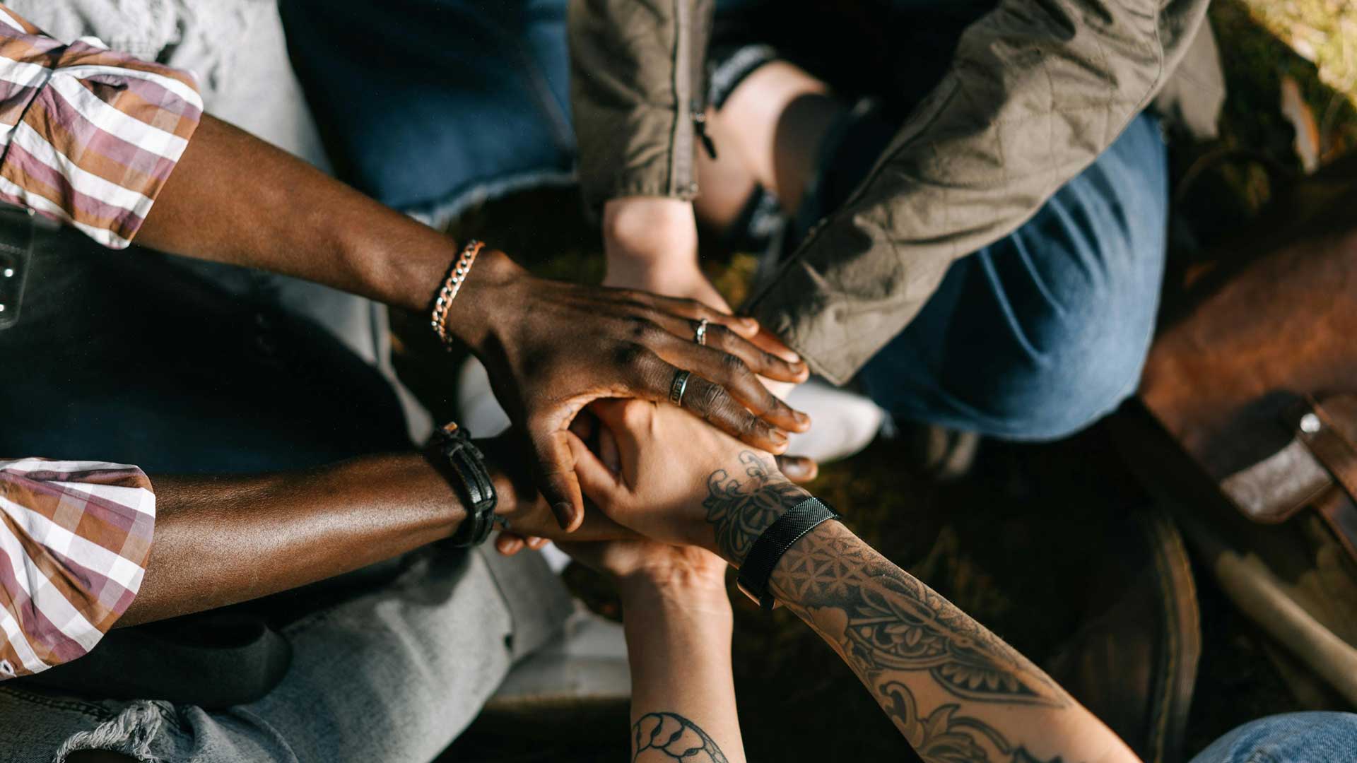 Quatre mains diverses empilées les unes sur les autres. Les mains présentent différentes couleurs de peau et des accessoires tels que des bracelets et des bagues, un bras étant tatoué.