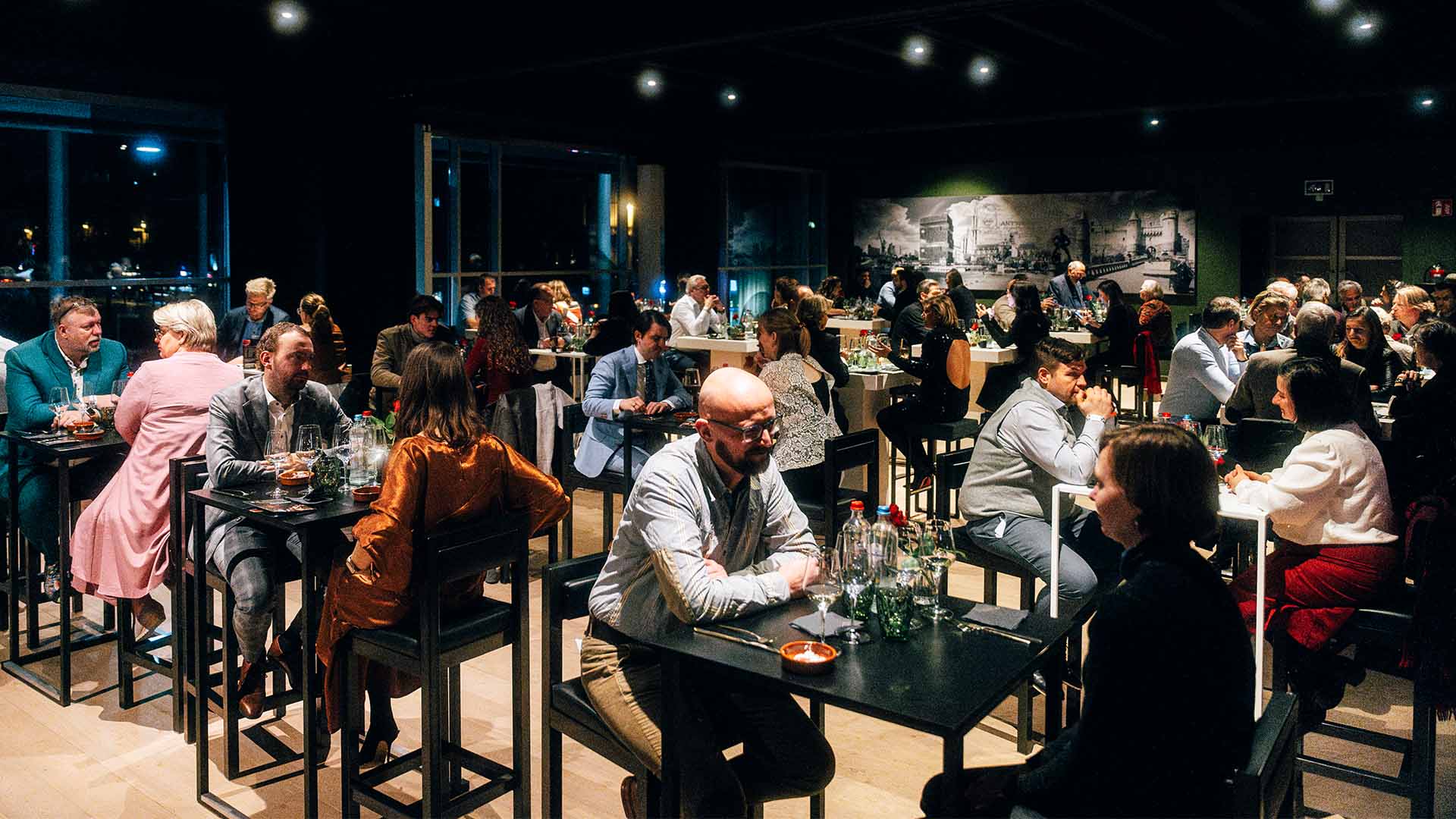 Overzicht van een drukbezochte evenementenruimte met gasten die aan hoge tafels zitten en met elkaar praten.