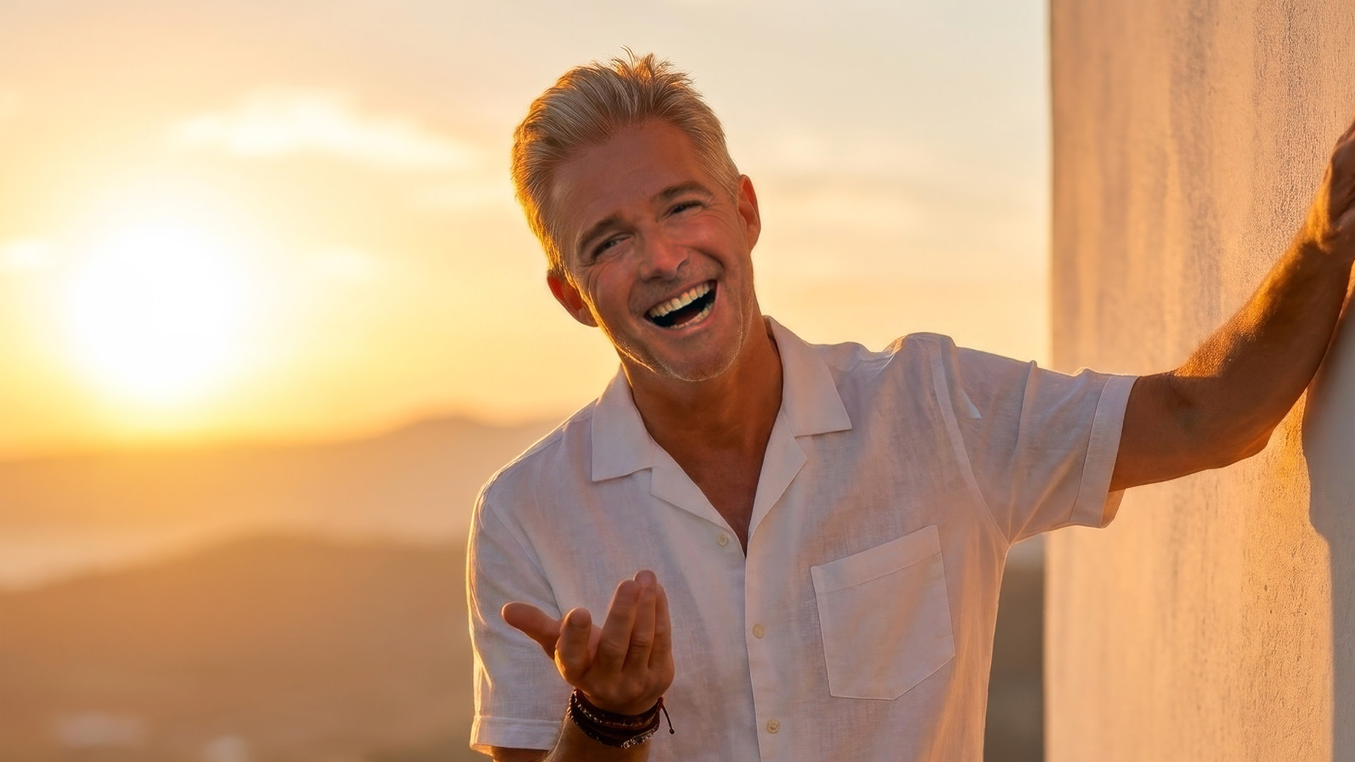 Christoff poseert lachend in een wit hemd tijdens zonsondergang, met een warme gouden gloed op de achtergrond.