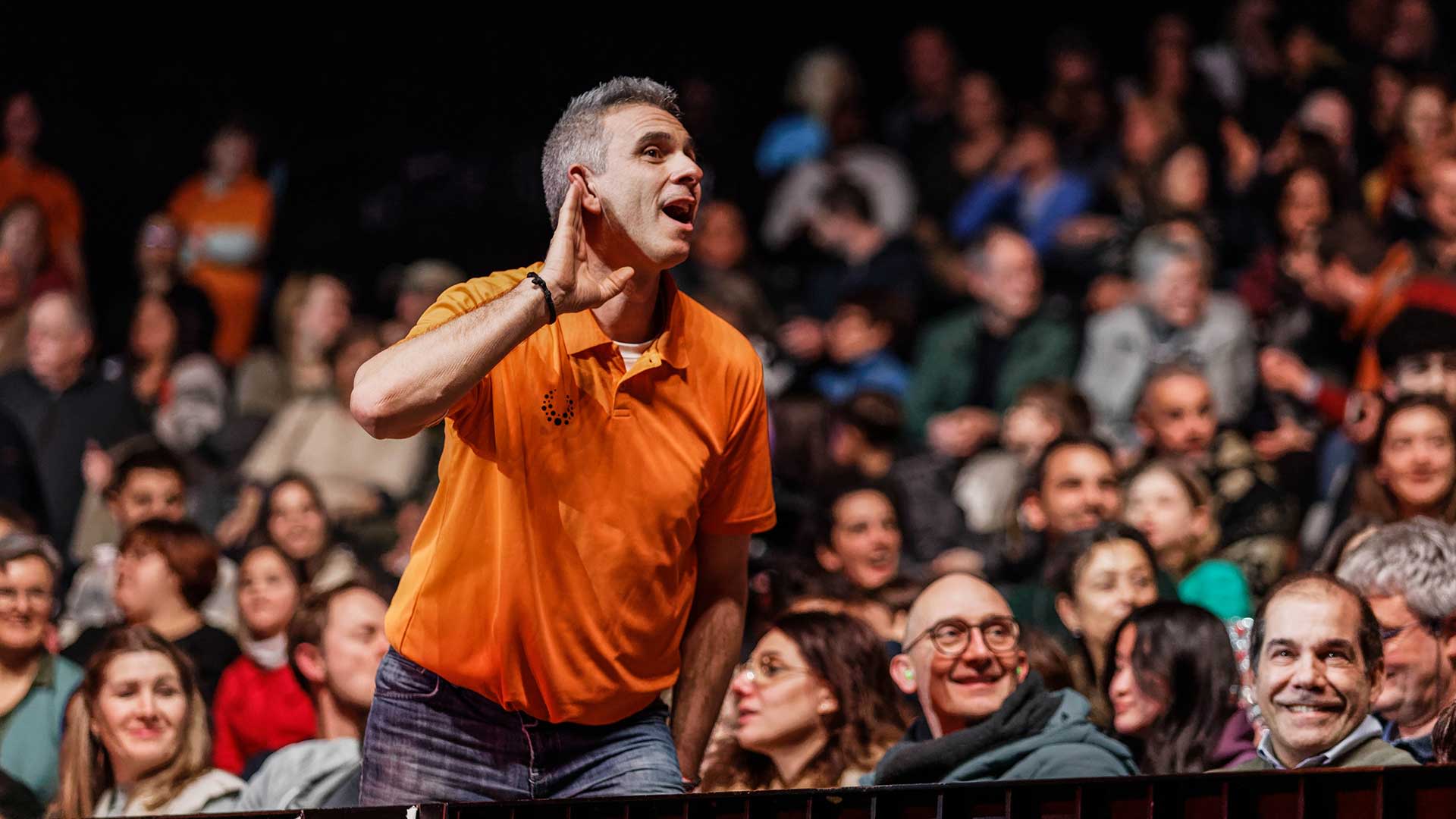 A man in an orange short-sleeved polo shirt and jeans stands in a crowd, with his hand cupped behind his ear as if listening, and looks up with a smile. He is in the foreground, and many other people are vaguely visible in the background, watching.