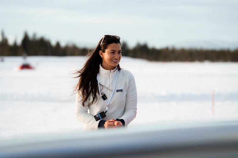 Woman with brown hair, white shirt and sunglasses smiling in front of a snowy landscape in the sunlight.