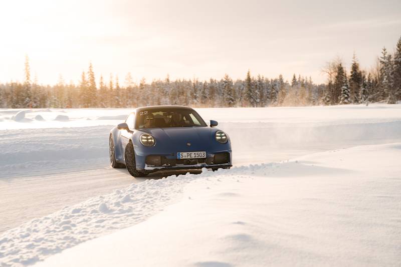 Blue Porsche 911 driving dynamically on a snowy road through a winter forest at sunrise.