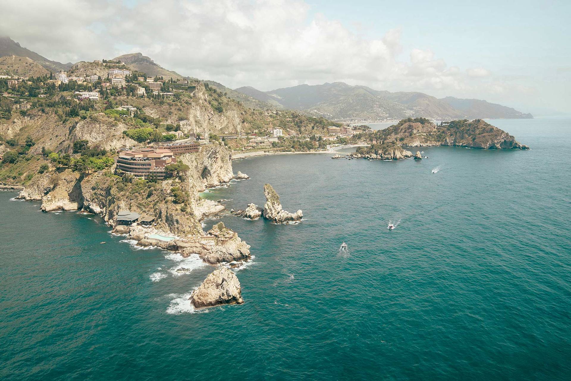 Rocky cliff coastline with buildings, boats at sea, and scattered clouds.