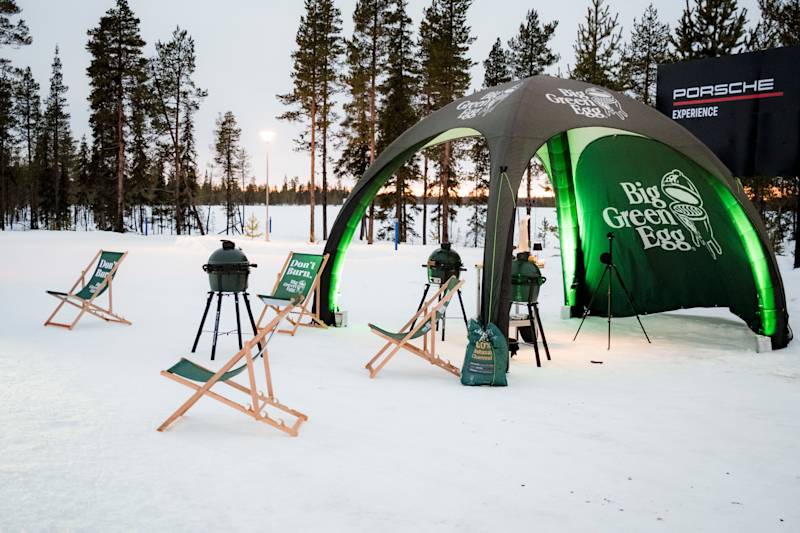 Big Green Egg grills and deck chairs under pavilion in snowy sunset setting, Porsche Experience sign in the background.