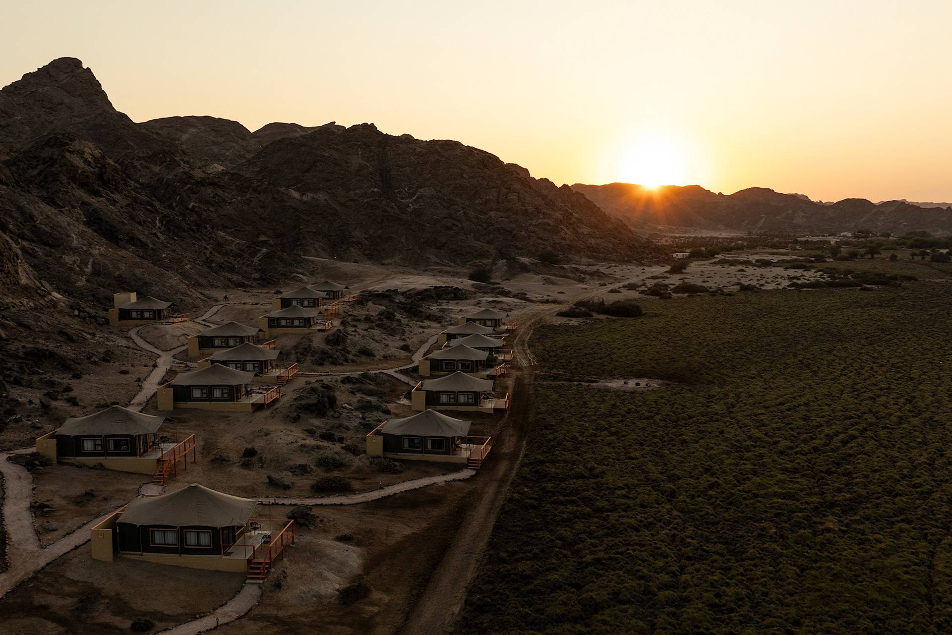 Desert camp with tented cabins at sunset, rocky terrain, and distant mountains.