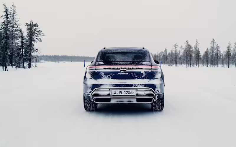 Rear view of a Porsche Macan Electric on a snow-covered road between pine trees under a grey sky.