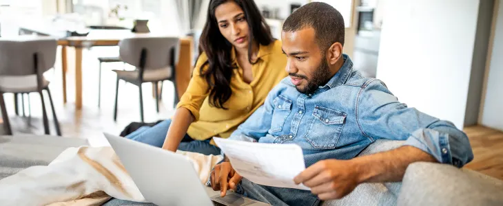 The image shows a couple sitting on a couch, reviewing documents together while using a laptop.