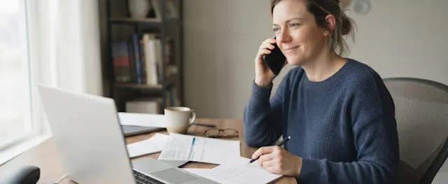 A woman working from home, talking on the phone while reviewing documents at her desk.