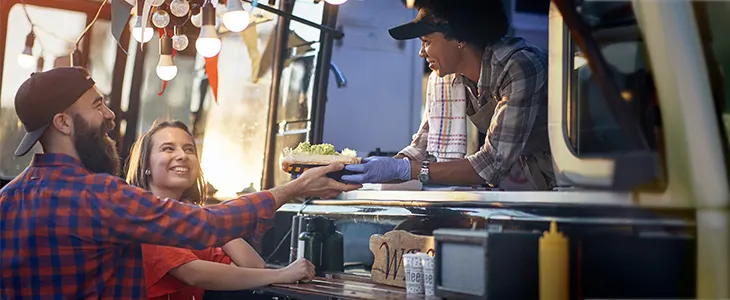 A cheerful couple receives food from a friendly vendor at a brightly lit food truck during an evening outing.