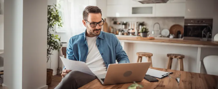 A man working remotely at home, focused on his laptop and reviewing documents at a kitchen table.