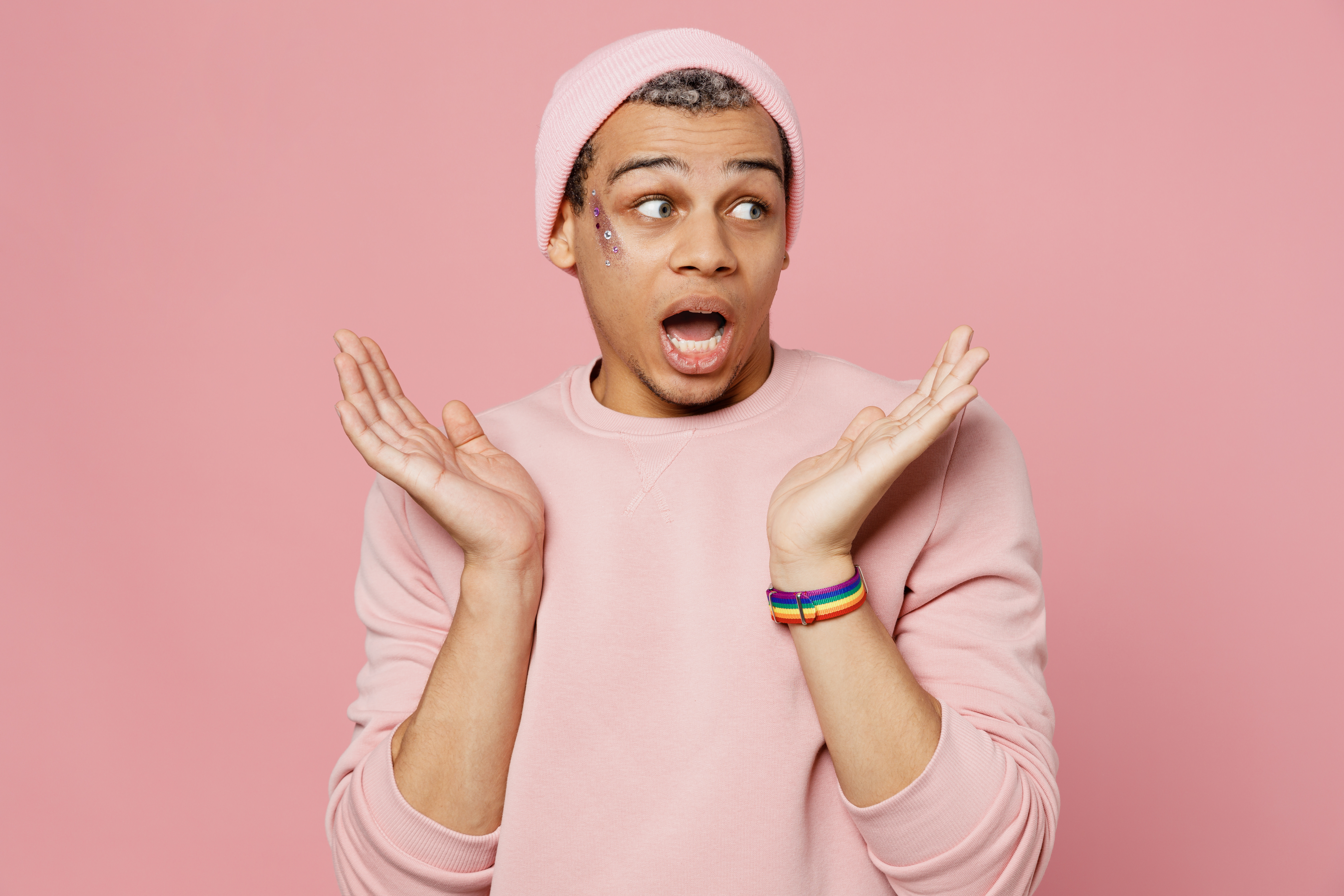 Young surprised shocked astonished gay man wear sweatshirt hat look on workspace area spread hands isolated on plain pastel light pink color background studio portrait