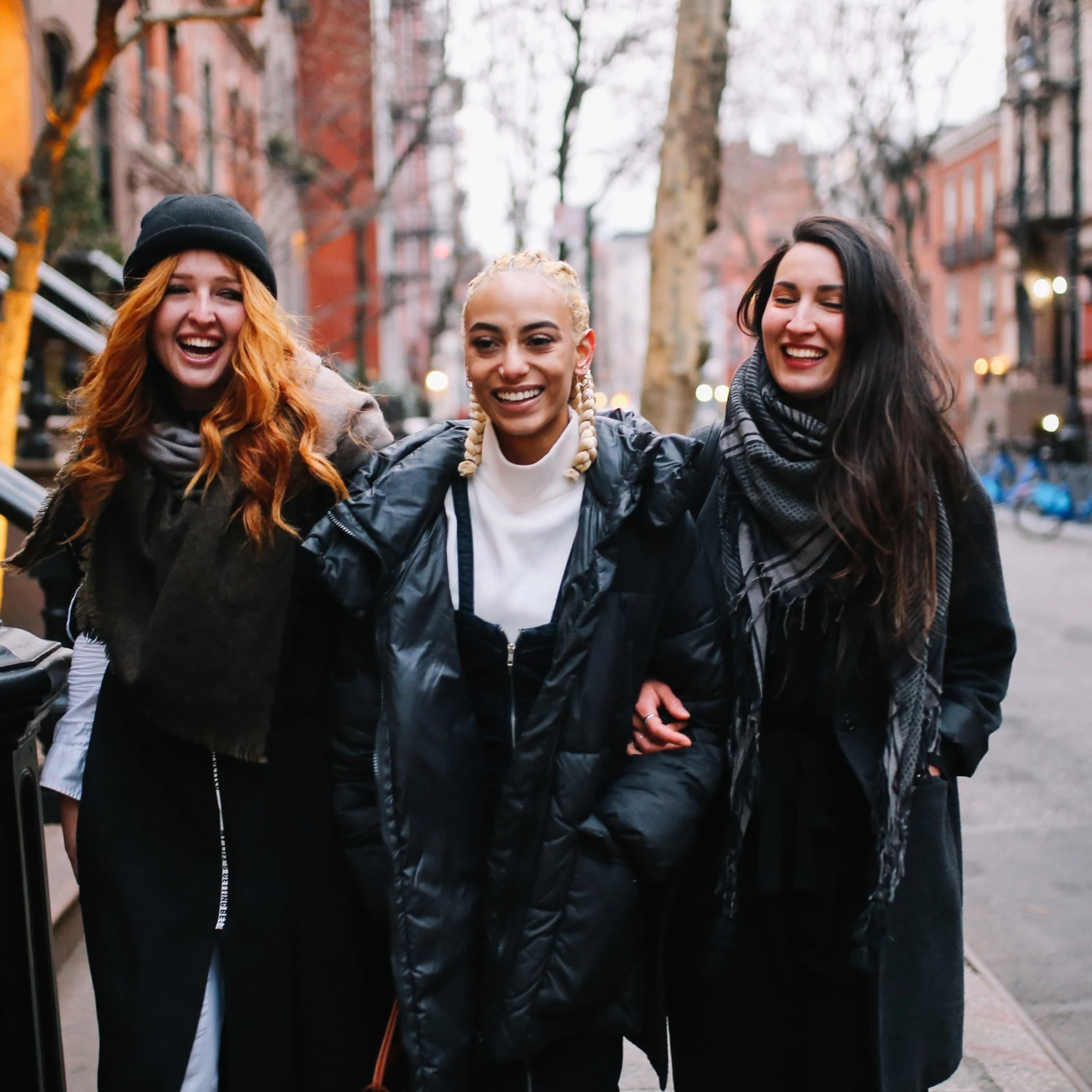 Three women walk with their arms linked on a city sidewalk 