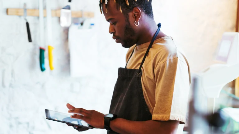 A Black person wearing a food service apron, tapping on a tablet device.
