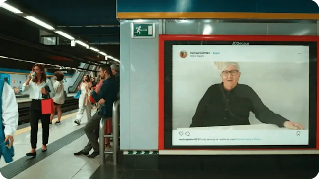 People wait for a train next to an ad for JCDecaux. The ad shows an elderly woman's social media post, featuring her sitting at a table.