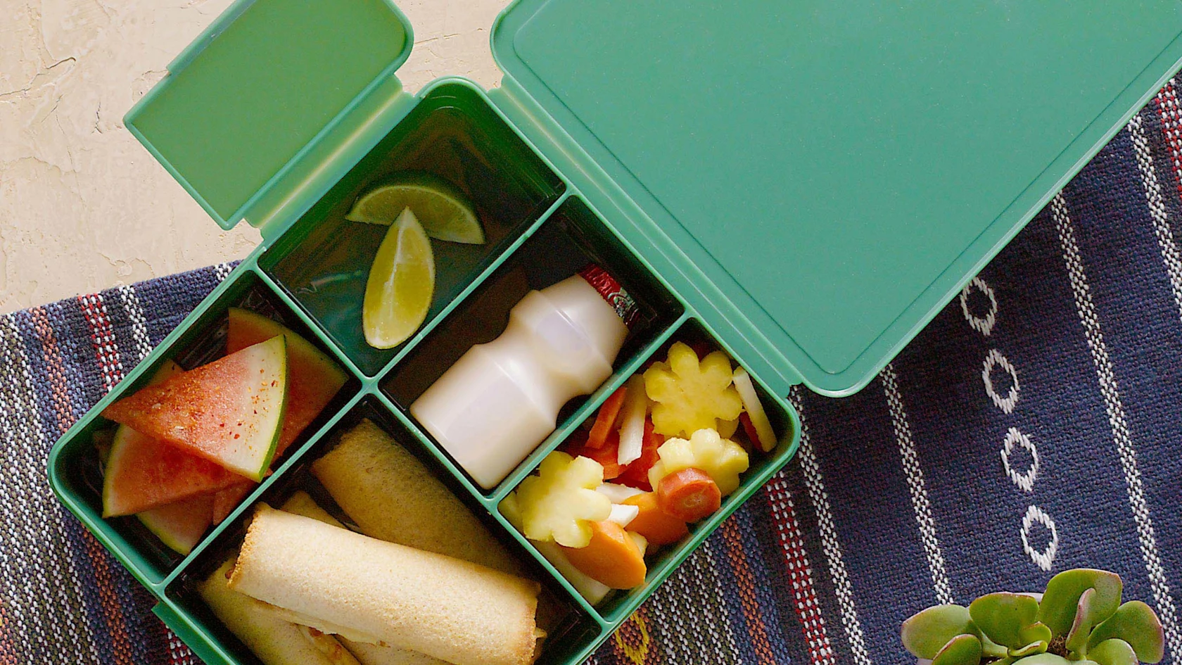 Green lunchbox with compartments holding watermelon slices, lime wedges, a drink bottle, spring rolls and cut vegetables on a tablecloth.