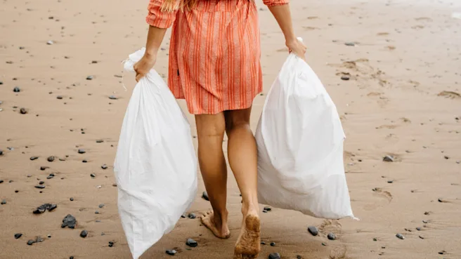 Une femme en robe orange marchant pieds nus sur une plage isolée et portant deux sacs poubelles blancs et pleins.