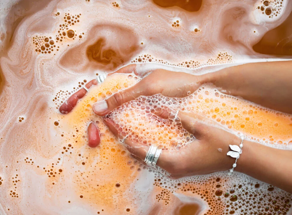 Une paire de mains dans une eau savonneuse orangée. La personne qui se lave les mains porte des bagues à chaque index et un bracelet à son poignet gauche.