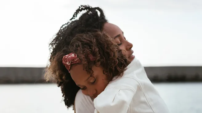 Mother and daughter wearing white dresses hugging in front of a body of water.