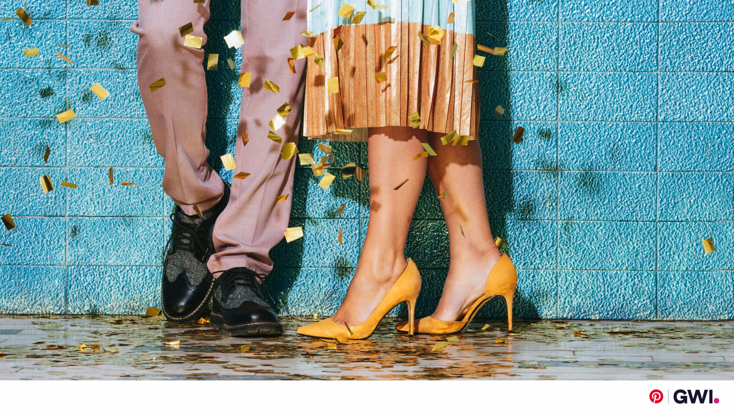 Pernas de duas pessoas próximas uma da outra contra uma parede azul, cercadas por confetes. A pessoa à esquerda veste uma calça na cor salmão e sapato social preto. A pessoa da direita usa uma saia bicolor e sapato de salto amarelo.