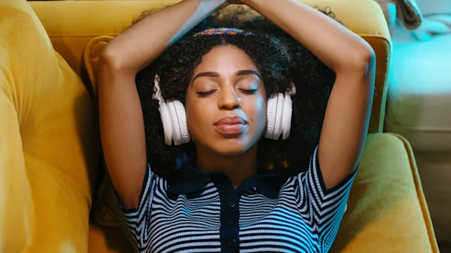 Black woman in a striped navy and light blue, short-sleeved top, eyes closed, wearing white headphones. She lies back on a soft yellow-orange couch, arms resting above her head.