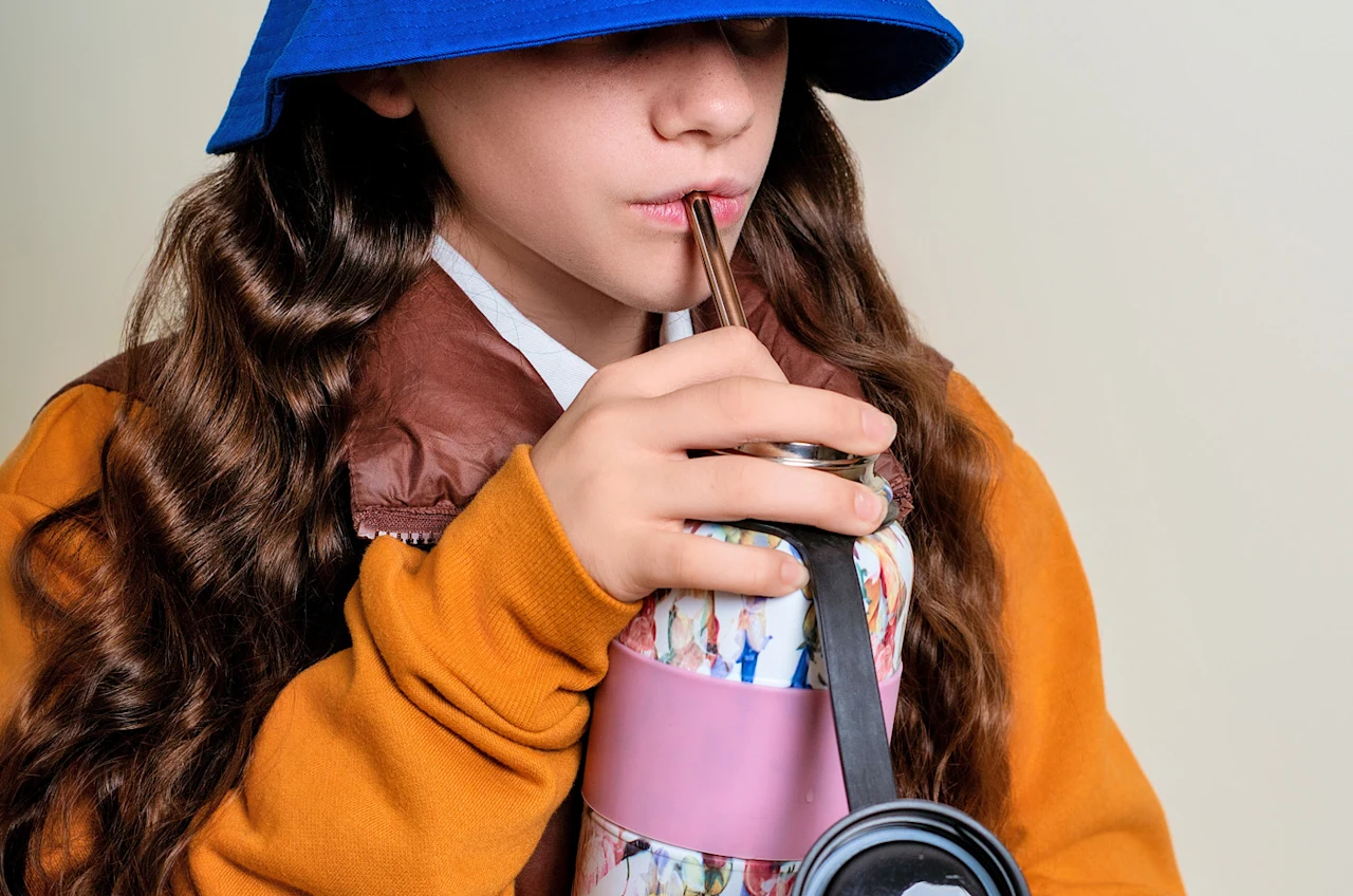 Child in a blue hat and orange jacket drinks from a reusable bottle with a metal straw, holding the bottle with both hands.