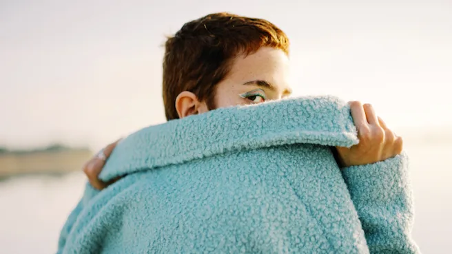 Woman with a brunette pixie cut and bright blue eyeliner wearing a fuzzy blue jacket peering over one shoulder